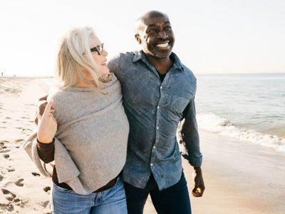 A mature Black man and White woman walk together, smiling, along a beach.