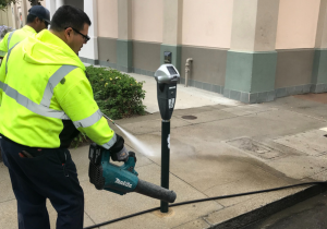 Two men: one pressure washing sidewalk, the other using a leaf blower to control flow of water towards vacuum