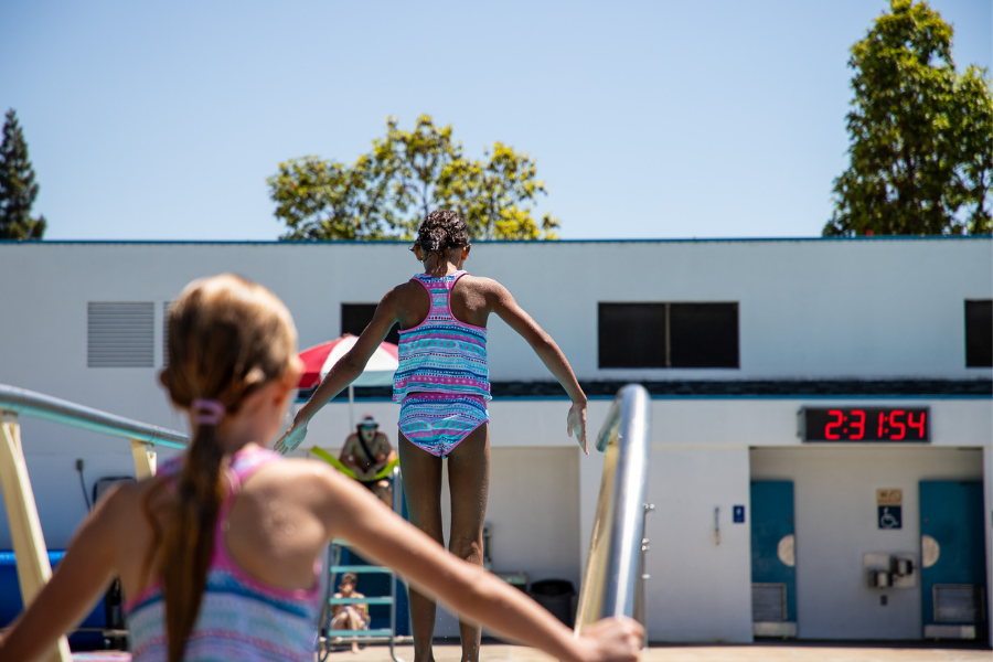 A picture of two youth standing on the diving board at the SLO Swim center, one getting ready to jump and one waiting on the stairs.