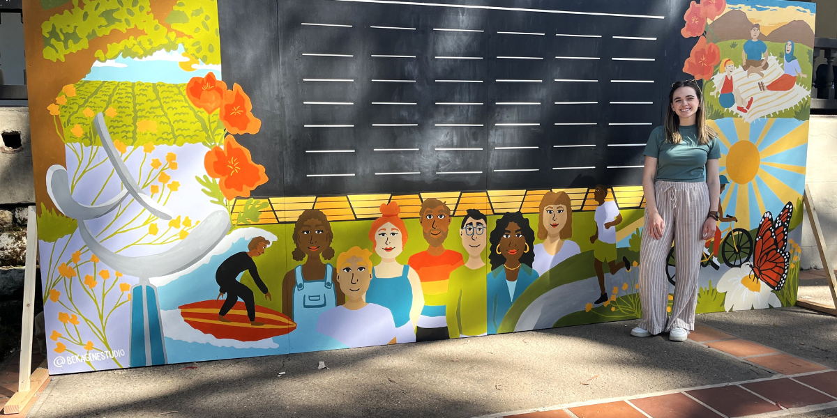 A picture of artist Rebekah standing next to the Community Mural in Mission Plaza in SLO